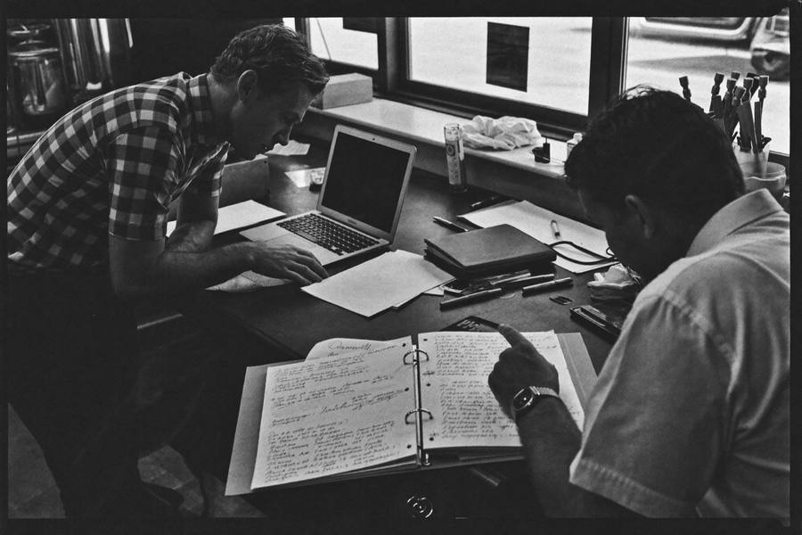 An estimator and operations manager leaning over a workbench, reviewing a technical schematic on a laptop beside an open binder of notes