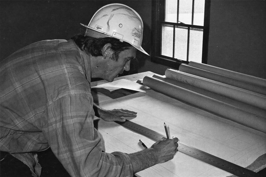 A construction estimator at a drafting table measuring a blueprint sheet with a scale ruler